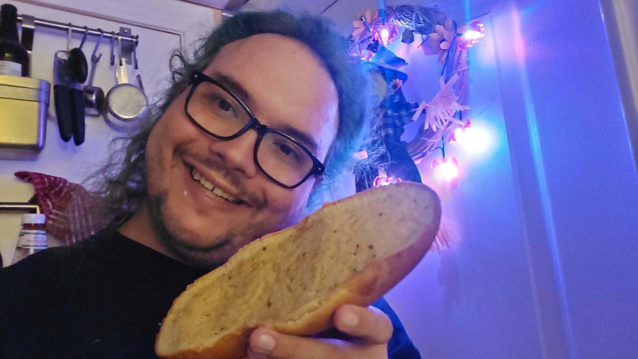 A man with long green hair and no beard holds a piece of garlic bread in a kitchen lit in purple light, Photo 1